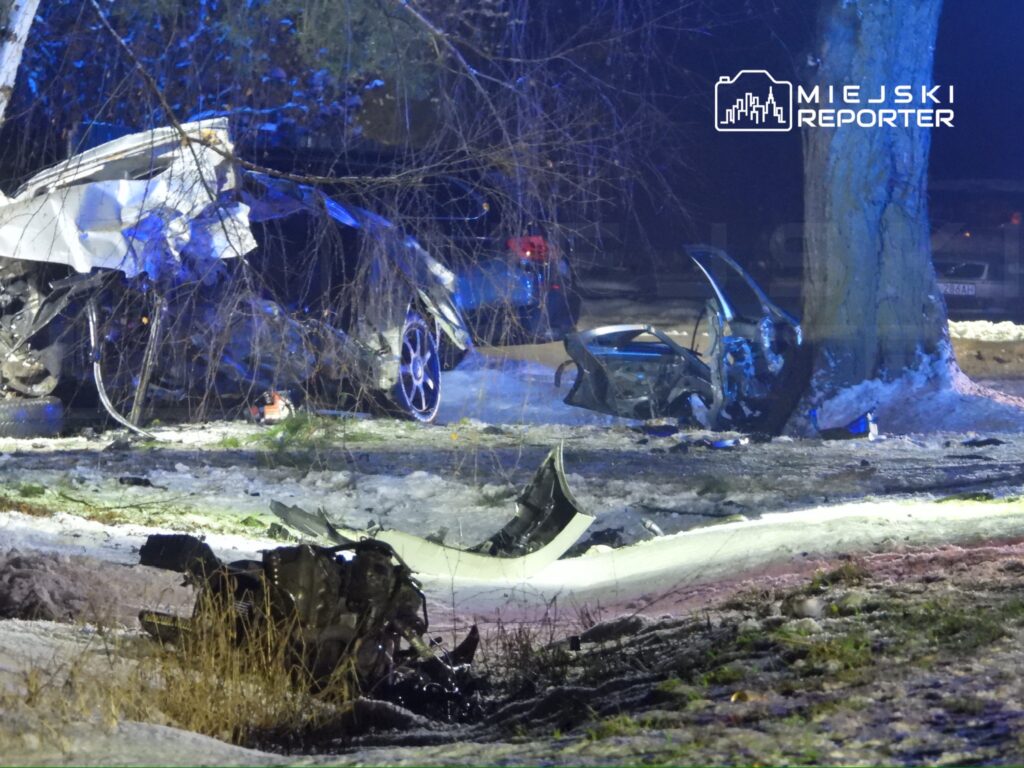 Zniszczony samochód osobowy rozbity o drzewo na zaśnieżonej drodze, obok leżące fragmenty pojazdu.
