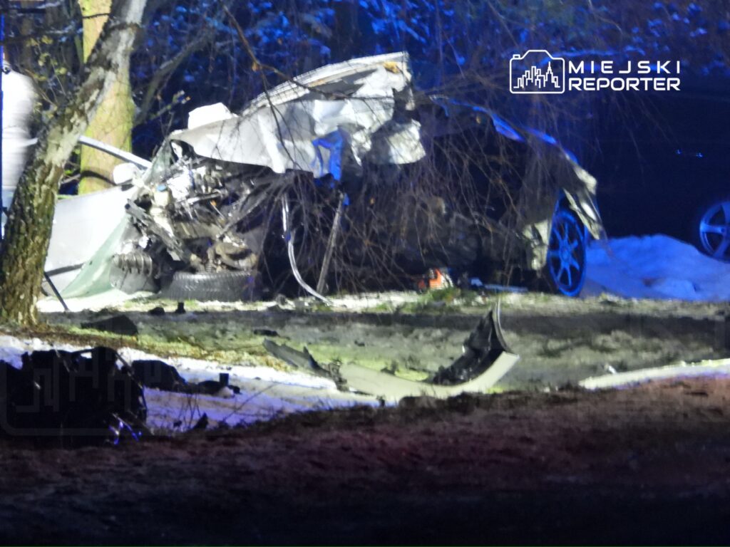 Zniszczony samochód osobowy leży na poboczu drogi, otoczony przez drzewa i śnieg, w nocy oświetlony niebieskimi światłami.