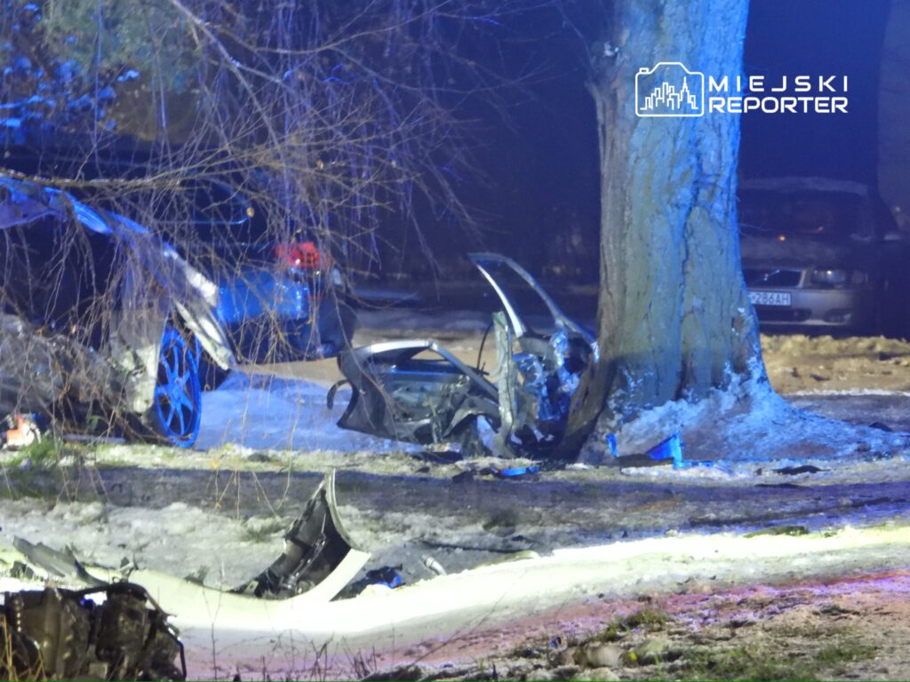 Zniszczony samochód osobowy leży przy drzewie na zaśnieżonej drodze, obok widać fragmenty karoserii i ślady opon.