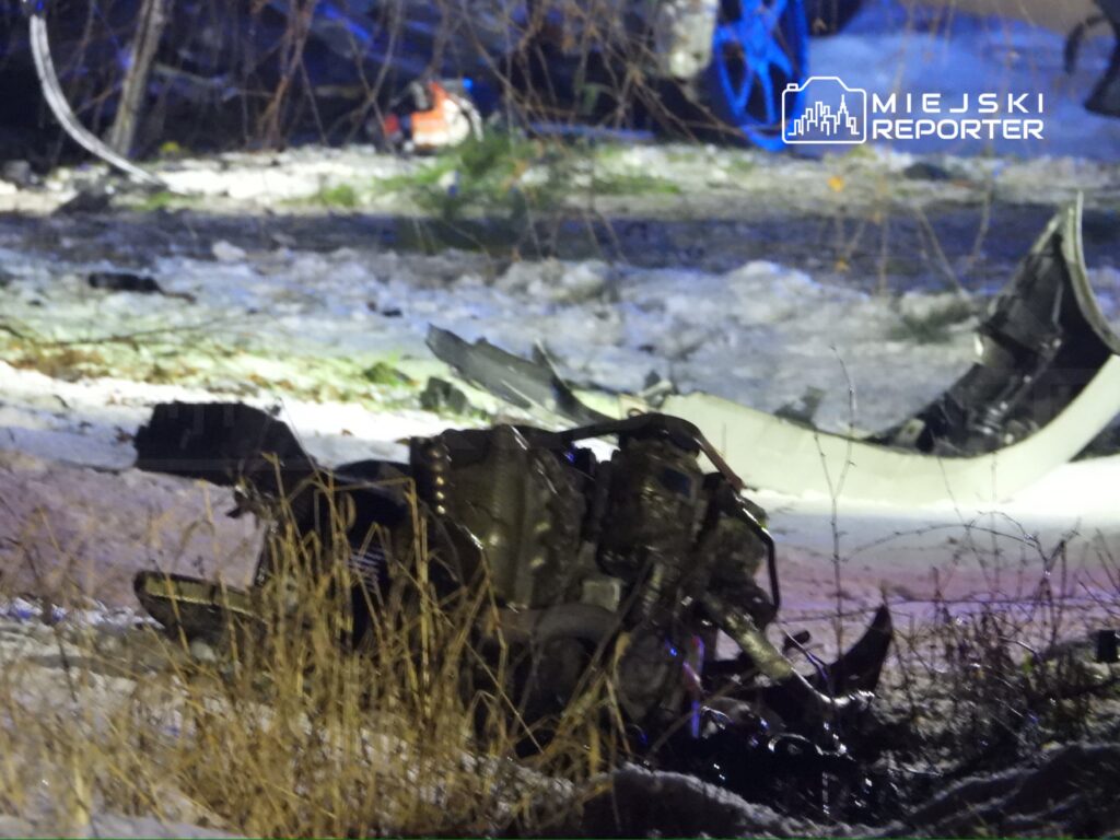 Zniszczony fragment pojazdu leży na zaśnieżonym terenie, w tle widoczne są niebieskie światła służb ratunkowych.