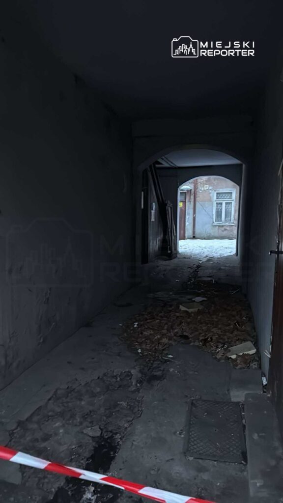 Wąski korytarz z widoczną warstwą liści i zniszczonym podłożem, prowadzący do oświetlonego wyjścia.