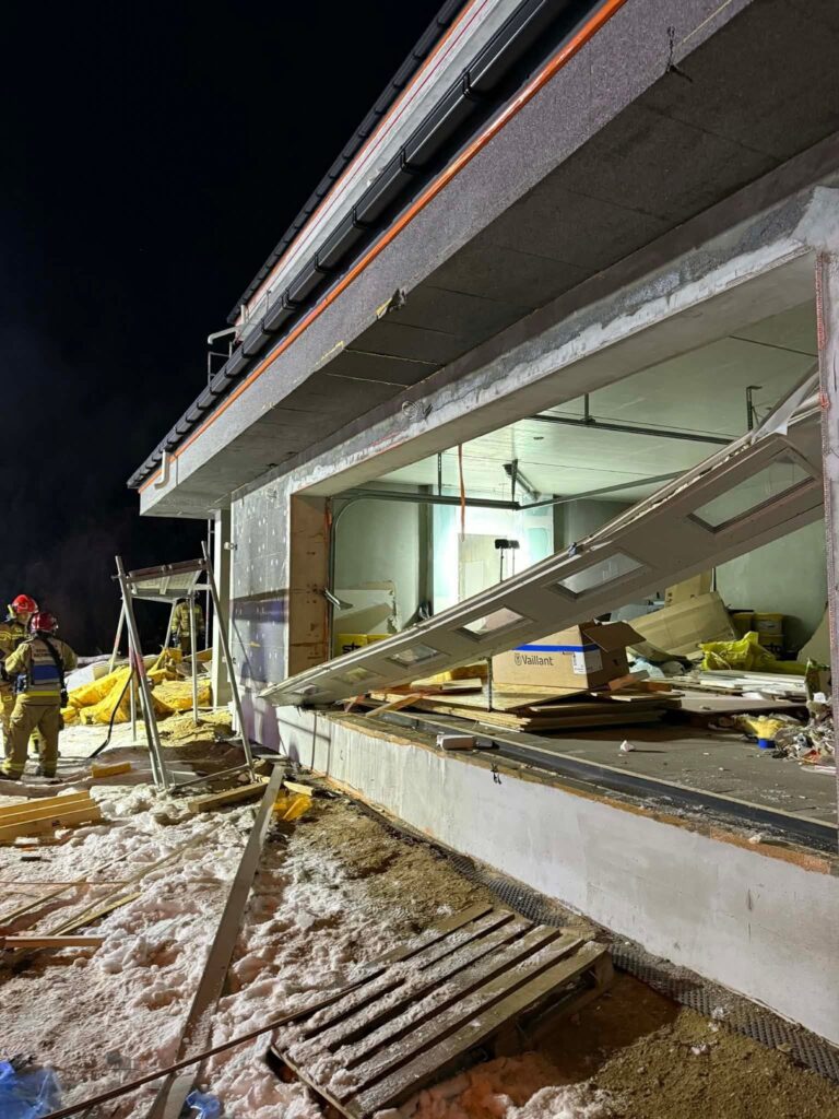 Grupa strażaków w mundurach obserwuje uszkodzoną elewację nowo budowanego obiektu w nocy. W tle widać załadunek materiałów budowlanych.