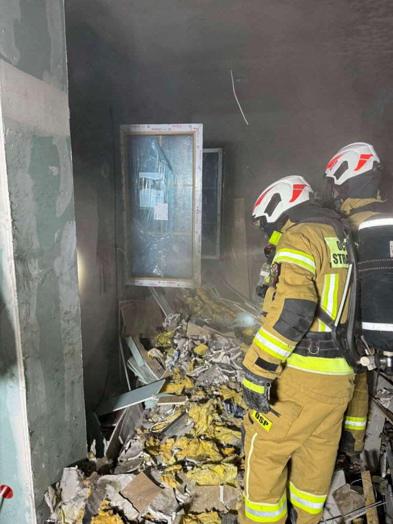 Strażacy w sprzęcie ochronnym przeszukują zniszczone pomieszczenie wypełnione gruzem i dymem.
