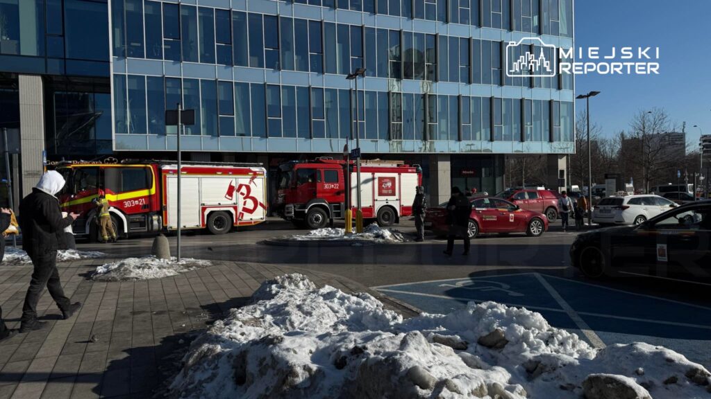 Strażacy przy wozach strażackich interweniują przed nowoczesnym biurowcem, obok leży śnieg.