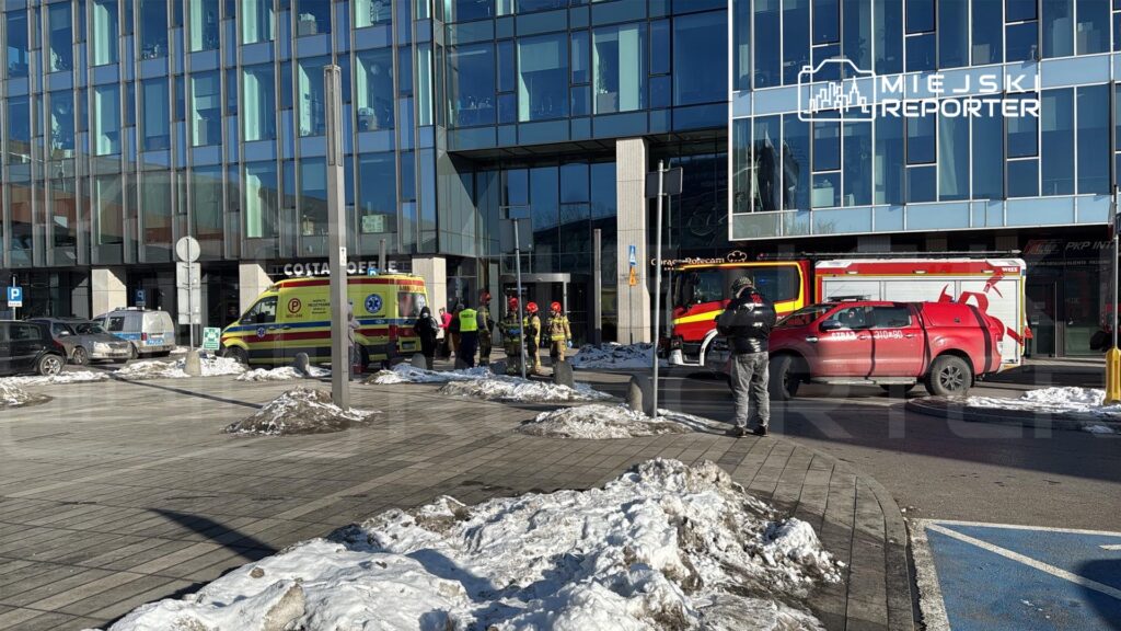 Zespół ratownictwa medycznego oraz strażacy interweniują przed nowoczesnym biurowcem, obok parkujących samochodów.