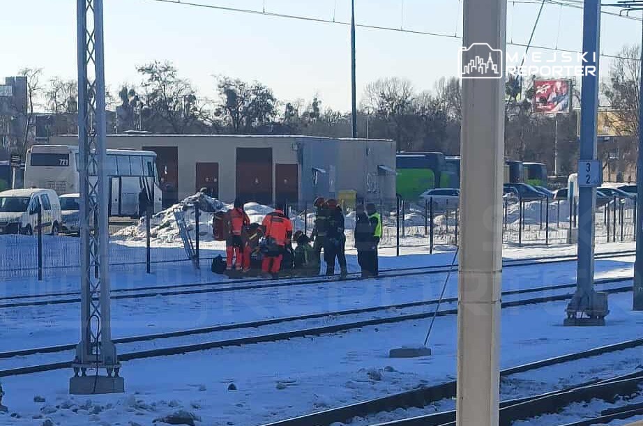 Grupa ratowników w pomarańczowych kombinezonach udziela pomocy na torach kolejowych w zimowym otoczeniu.