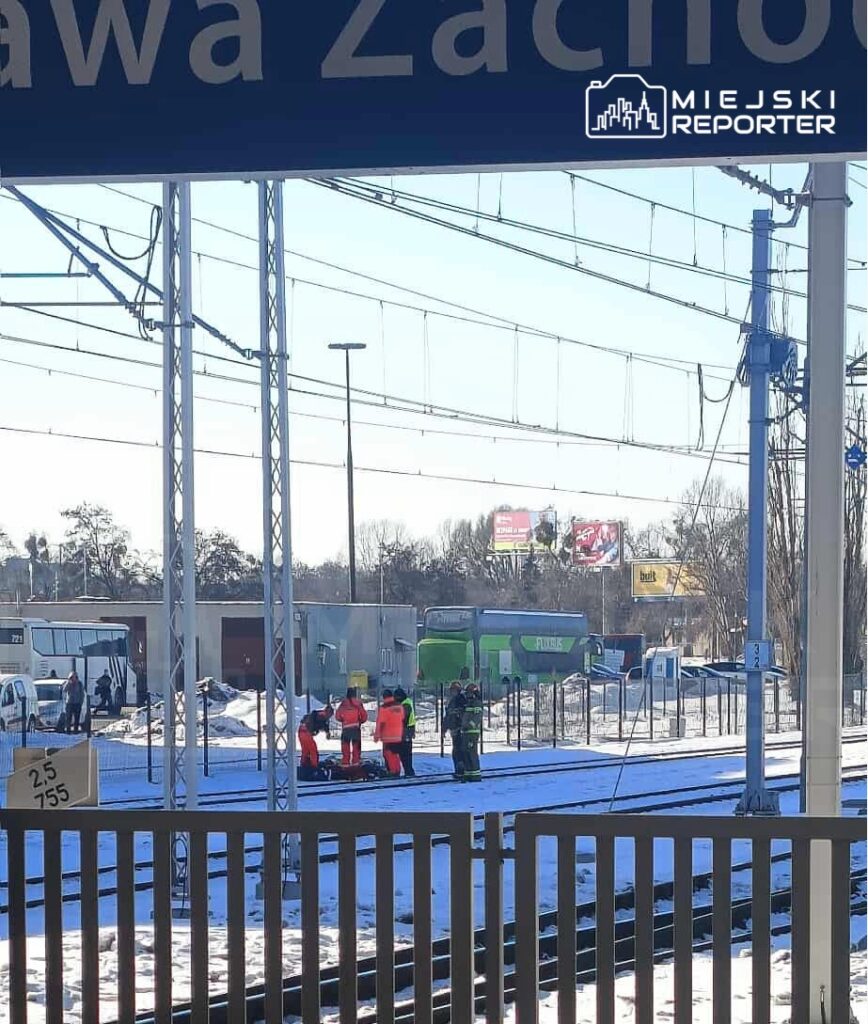 Grupa ratowników w pomarańczowych kamizelkach pracuje na torach kolejowych w zimowym otoczeniu stacji.