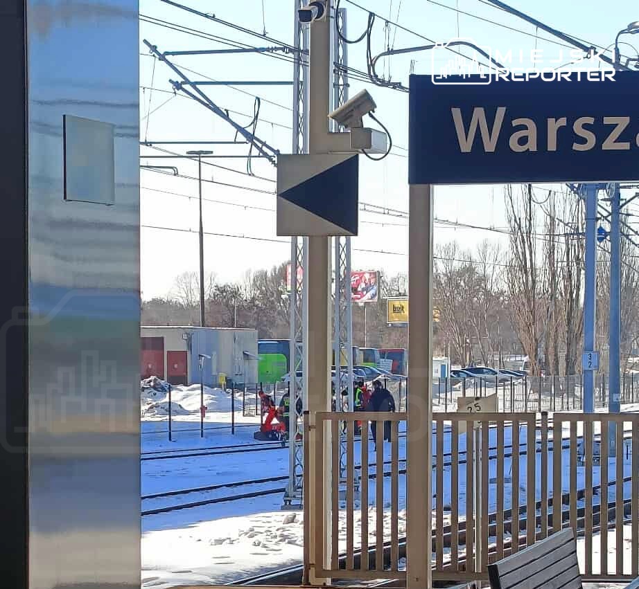 Na stacji kolejowej, znak z nazwą "Warszawa" oraz kamery monitorujące, w tle prace na torach w zimowej scenerii.