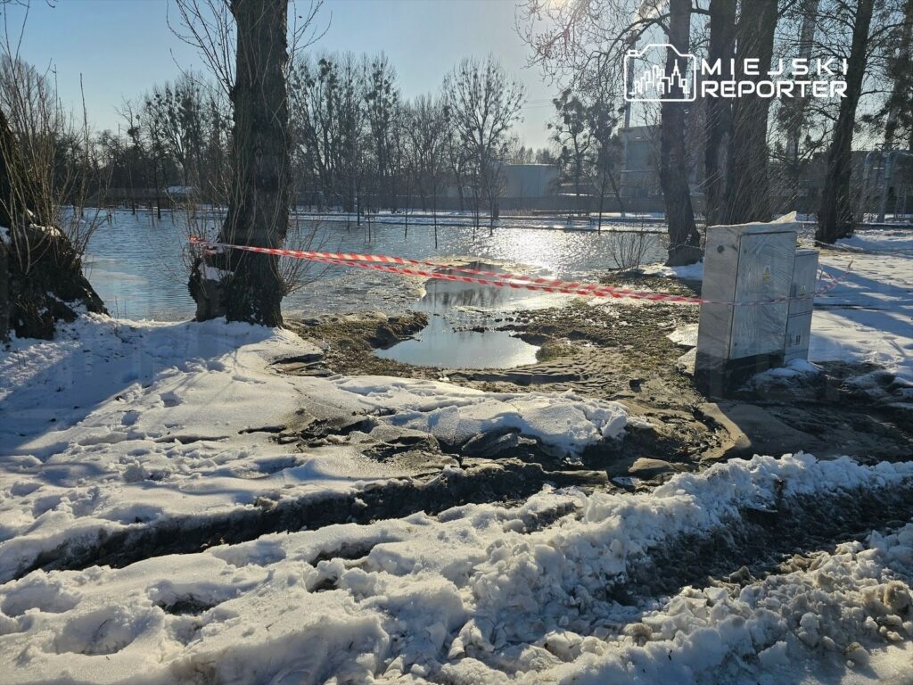 Zabezpieczony teren z uszkodzoną nawierzchnią przy zamarzniętym zbiorniku wodnym, otoczony śniegiem i drzewami.