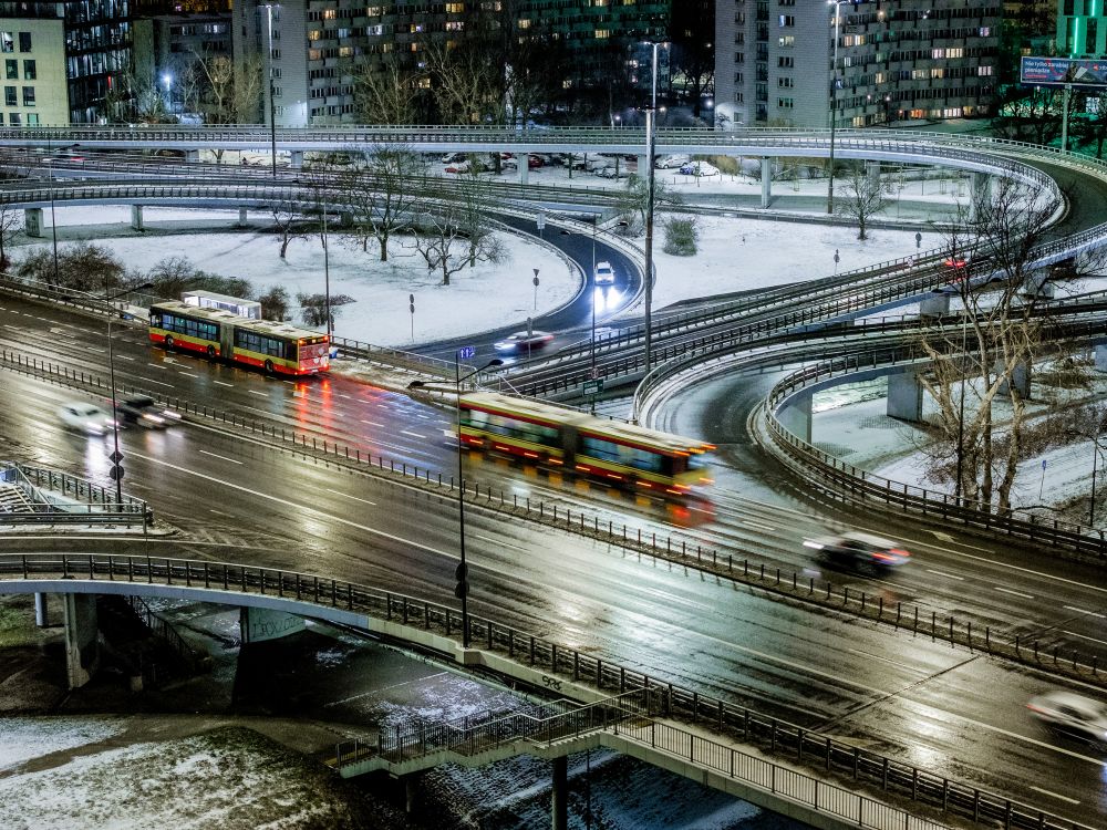 Ruchliwe skrzyżowanie z autobusami i samochodami, pokryte śniegiem, w nocy z oświetleniem miejskim.