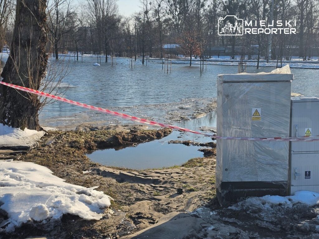 Zasypane wodą tereny zielone z wystającymi drzewami, w tle widoczna skrzynka elektryczna owinięta folią.