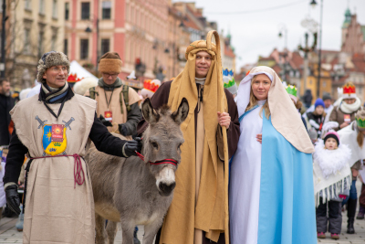 Grupa osób w kostiumach historycznych, w tym mężczyzna z osłem, idzie ulicą podczas parady bożonarodzeniowej.