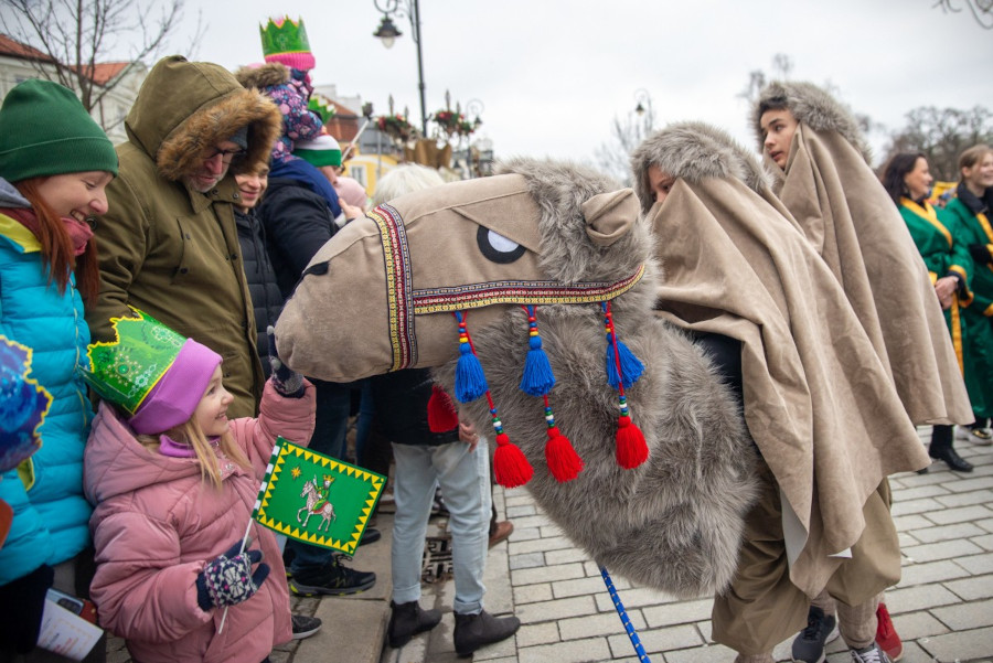 Grupa dzieci i dorosłych w kolorowych strojach bawi się podczas parady, a jedno z dzieci trzyma kartkę z rysunkiem.