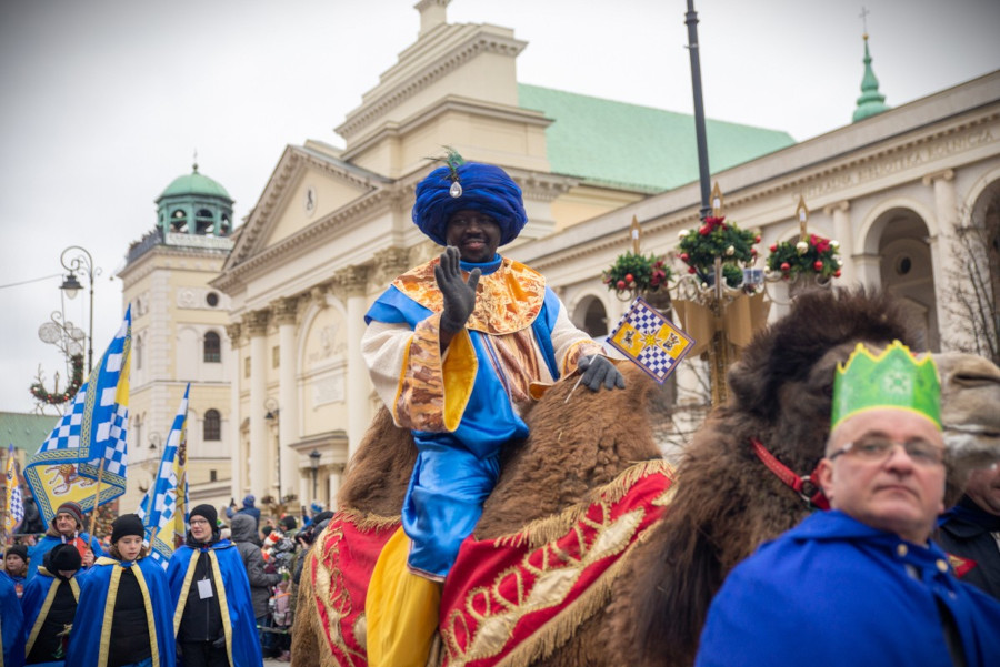 Mężczyzna w kolorowym stroju, siedzący na wielbłądzie, macha ręką podczas parady w miejskim otoczeniu.