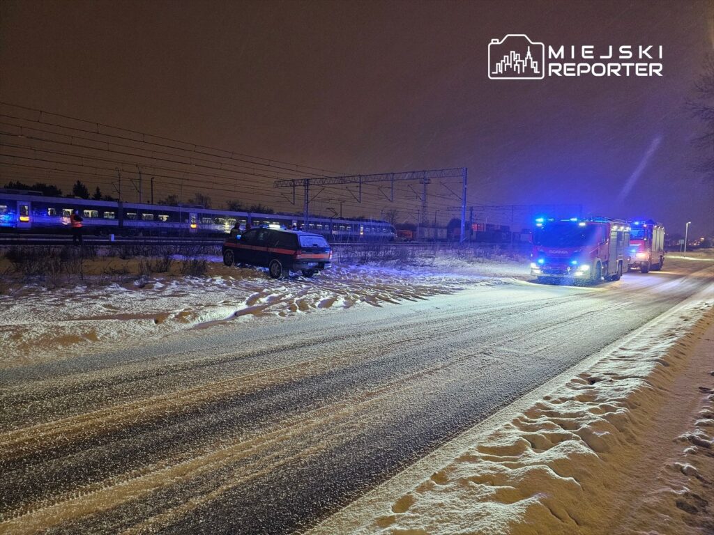 Samochód stoi na poboczu drogi w zaśnieżonym terenie, obok przejeżdża pociąg, a w tle widoczne są pojazdy straży pożarnej z włączonymi sygnałami.