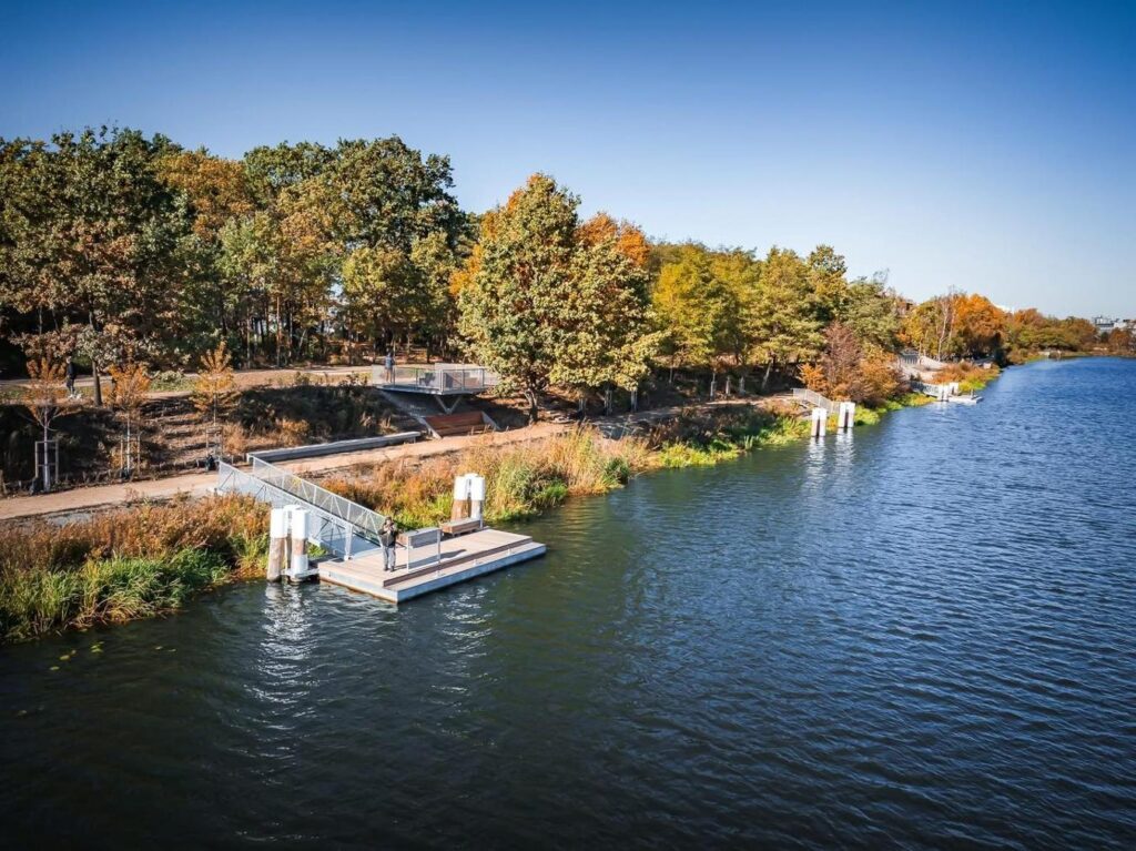 Mężczyzna stojący na pomostku nad rzeką, otoczony kolorowymi drzewami w jesiennej scenerii.