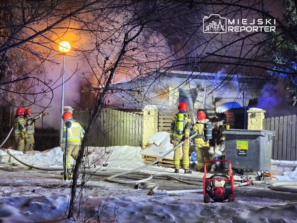 Grupa strażaków w hełmach i specjalnych ubraniach gasi pożar w pobliżu ogrodzenia, w otoczeniu śniegu i dymu.