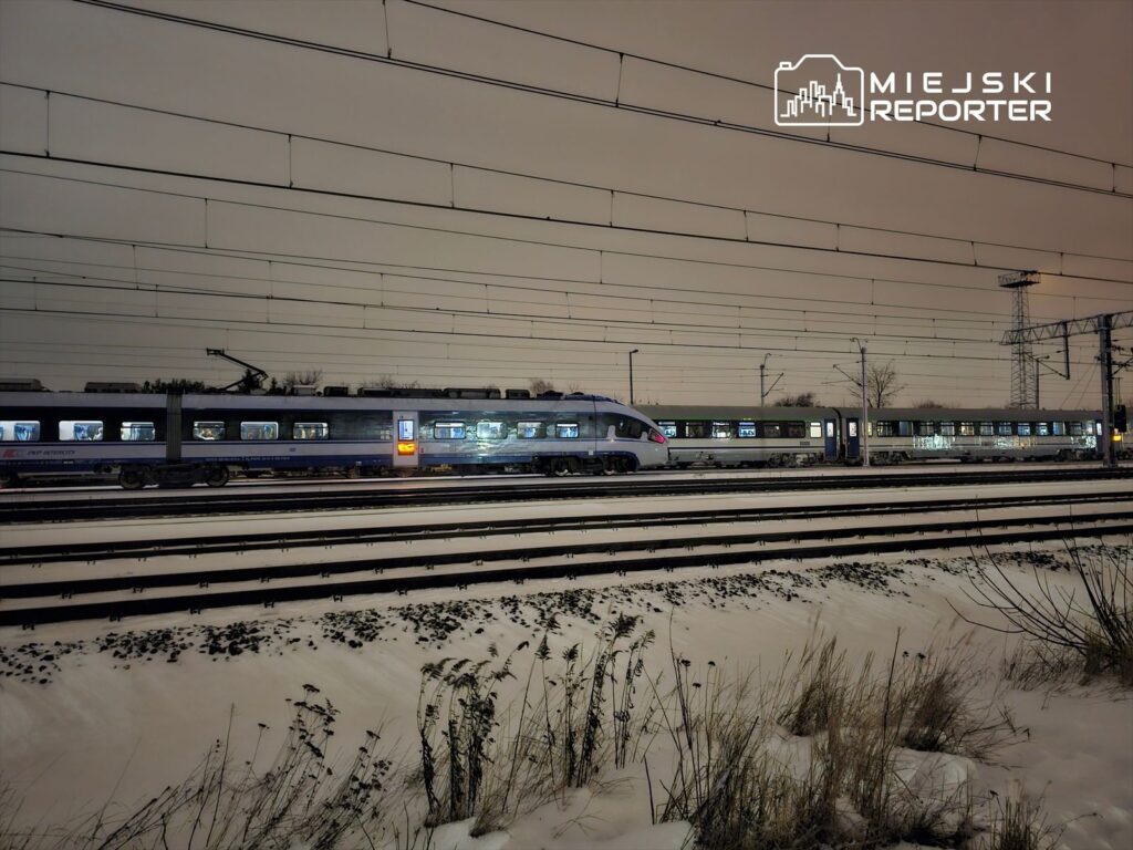 Pociąg osobowy zatrzymany na zaśnieżonym torze, z oświetlonymi oknami w nocy. W tle widoczne linie kolejowe.