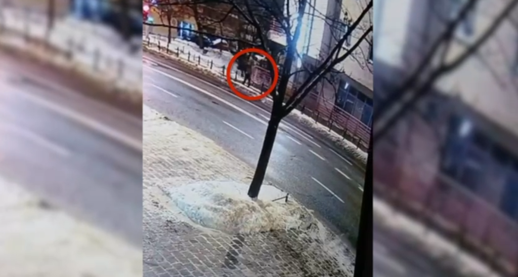 Grupa osób stojąca na chodniku w pobliżu ulicy, otoczona drzewami i budynkami w tle.