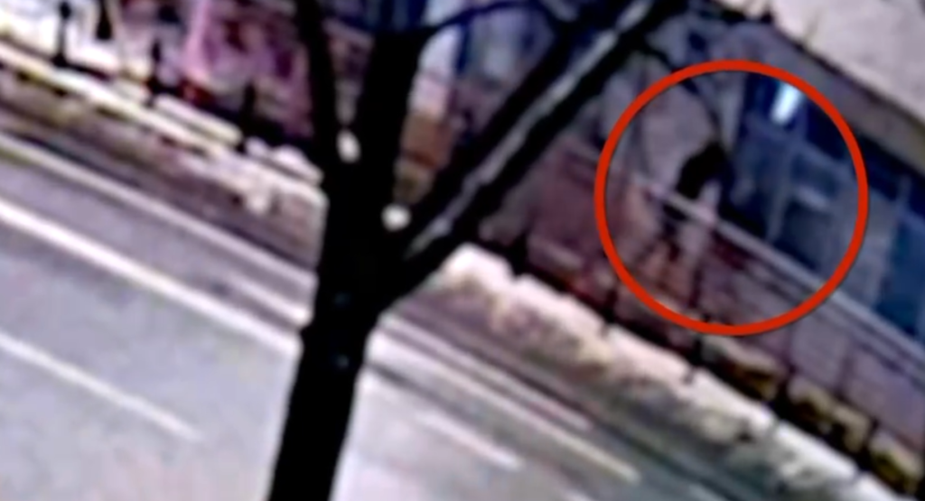 Czarna postać stojąca na barierce w pobliżu ulicy, w tle widoczne budynki i drzewa.