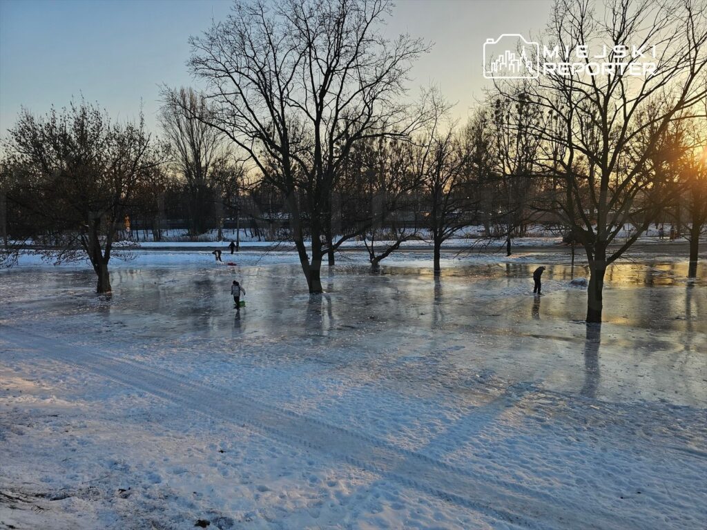 Dzieci bawią się na zamarzniętej powierzchni w parku, otoczone nagimi drzewami w blasku zachodzącego słońca.