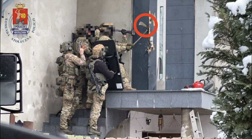 Grupa uzbrojonych policjantów w mundurach szturmowych używa narzędzia do wyważenia drzwi w budynku, otoczeni przez śnieg.