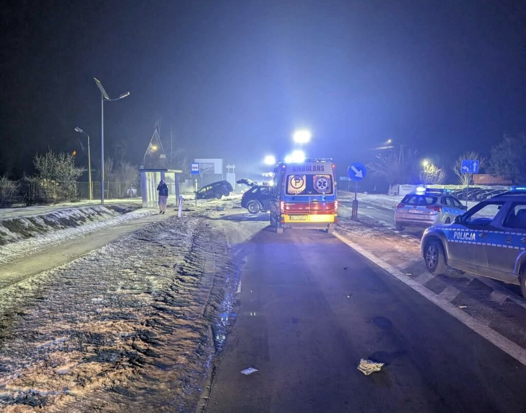 Na oświetlonej ulicy nocą, karetka pogotowia oraz kilka radiowozów policyjnych stoją obok uszkodzonego samochodu na poboczu.