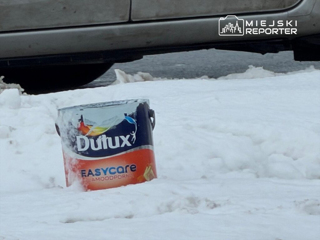 Pojemnik po farbie Dulux umieszczony na zaśnieżonym chodniku obok samochodu.