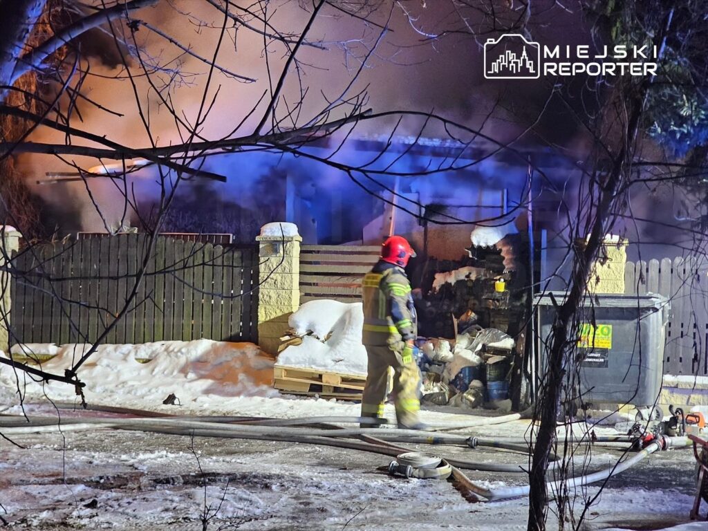 Strażak w hełmie bada miejsce pożaru w otoczeniu śniegu i drewnianego ogrodzenia. W tle widać dym.