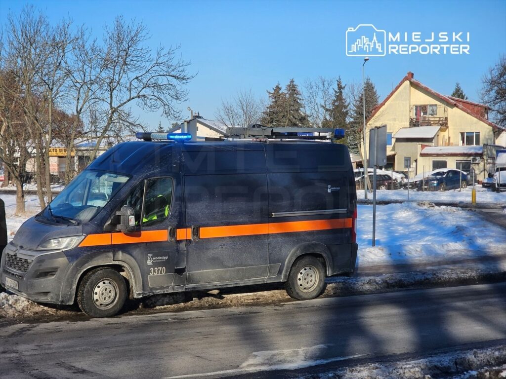 Funkcjonariusz w uniformie obserwujący teren obok radiowozu z niebieskim światłem na zaśnieżonej ulicy.
