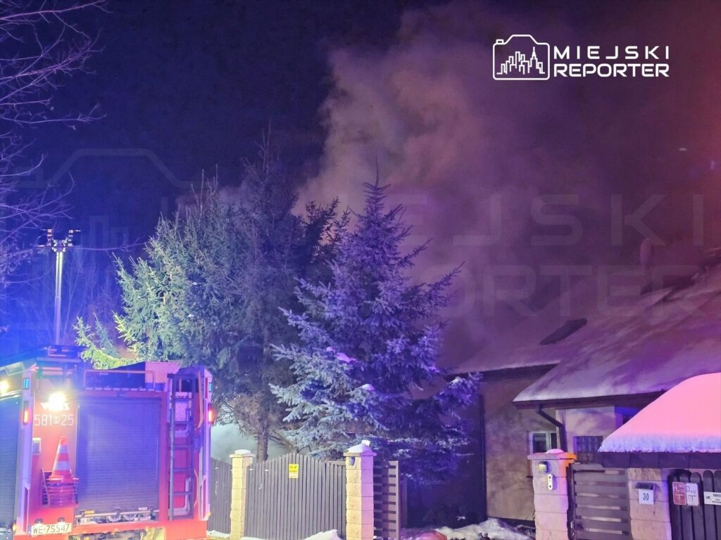 W nocy straż pożarna interweniuje przy pożarze budynku, otoczonego drzewami i ogrodzeniem, w tle widoczne dym i światła alarmowe.