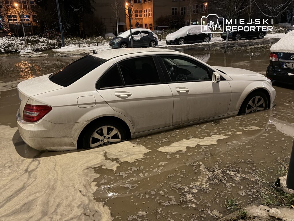 Biały samochód osobowy zanurzony w wodzie na zalanym parkingu, otoczony przez inne pojazdy i ośnieżone drzewa.