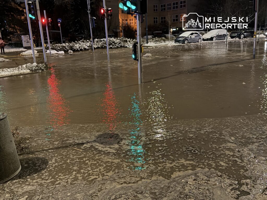 Zalany skrzyżowanie z odbiciem świateł drogowych w wodzie oraz zamarzniętymi samochodami w tle.