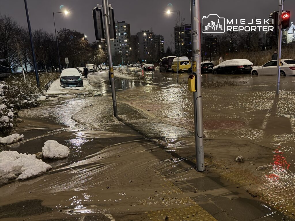 Zalany chodnik z widocznymi śladami błota, obok zaparkowane samochody pokryte śniegiem, nocna sceneria miejskiego osiedla.