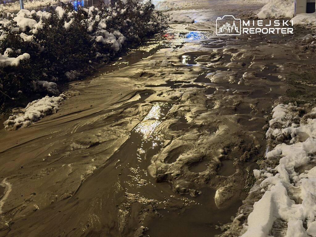 Mokry chodnik z błotem i śniegiem oraz odbiciem świateł miejskich w nocy.