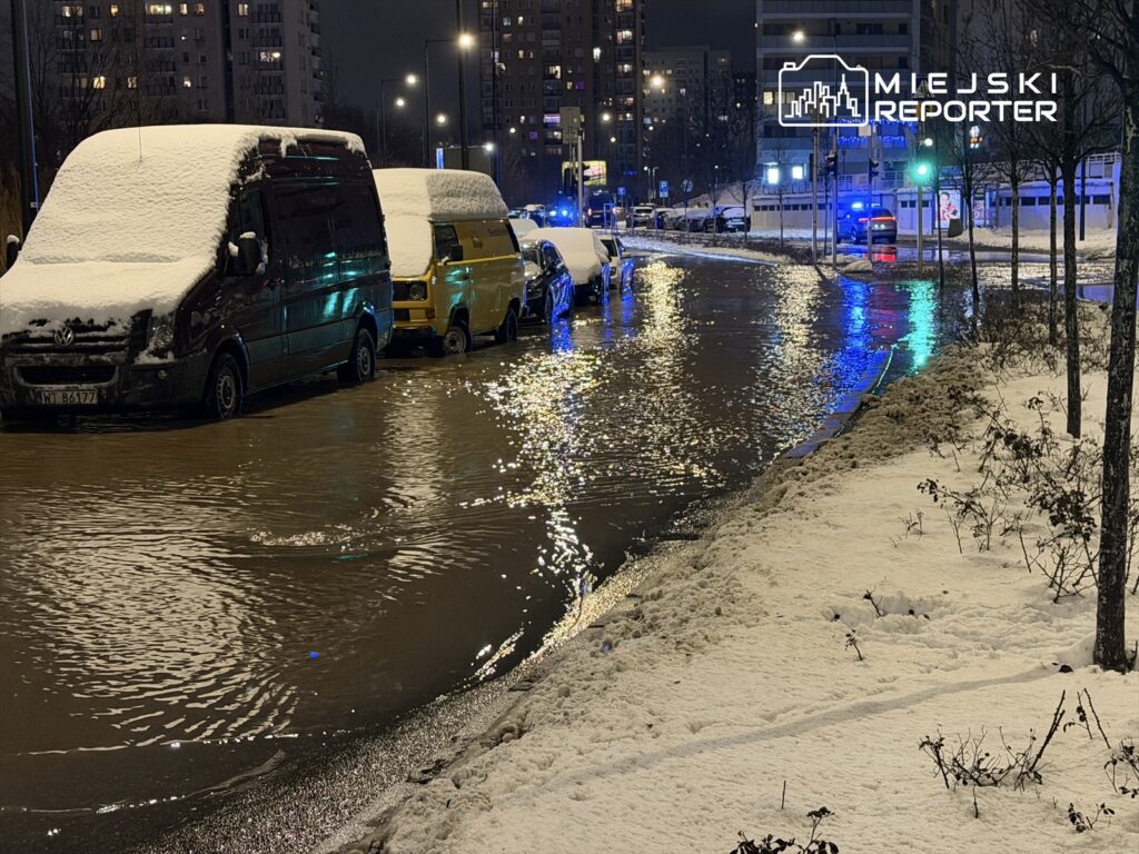 Zatopiona ulica w nocy, pokryta śniegiem, z zaparkowanymi samochodami i odbijającymi się światłami.