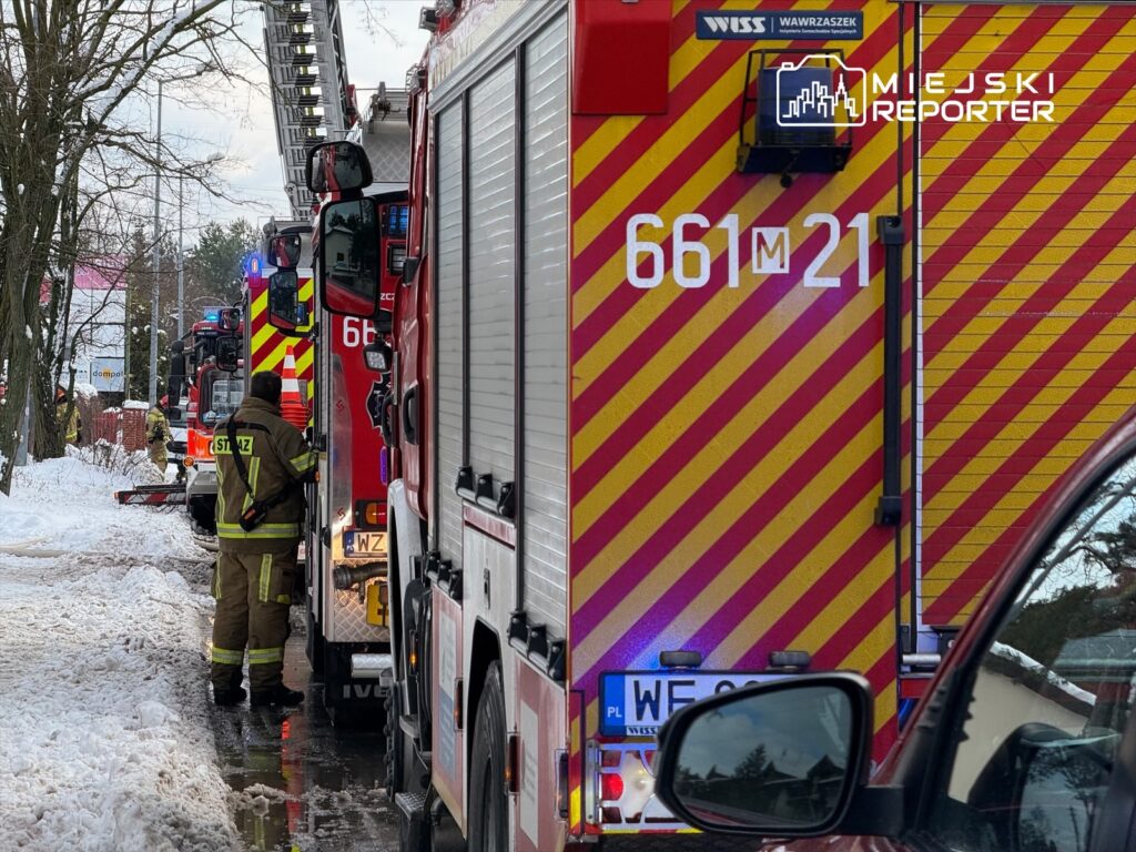 Strażak w mundurze stoi obok wozu strażackiego na zaśnieżonej ulicy, w tle widoczne inne pojazdy ratunkowe.