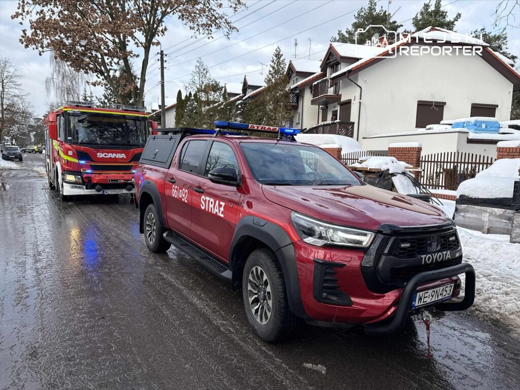 Czerwony samochód strażacki oraz pojazd ratunkowy zaparkowane na ośnieżonej ulicy w otoczeniu domów jednorodzinnych.