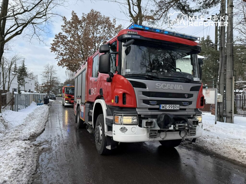 Czerwony wóz strażacki zaparkowany na zaśnieżonej ulicy, obok widoczne inne pojazdy ratunkowe.