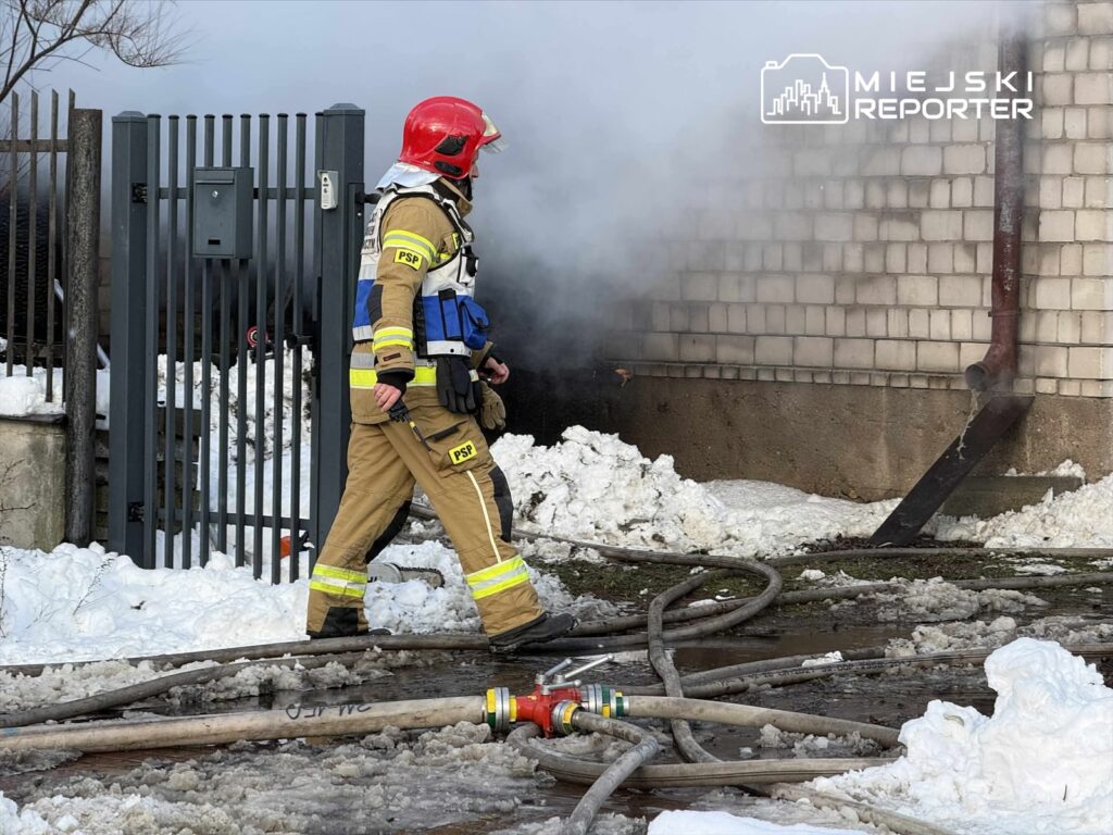 Strażak w hełmie i mundurze gaśniczym przemieszcza się obok oparów dymu w otoczeniu zaśnieżonej posesji.