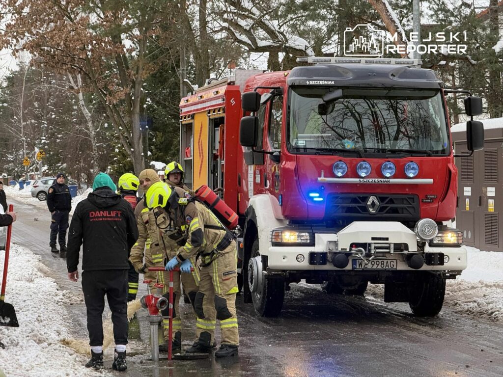 Grupa strażaków w kaskach i mundurach pracuje przy hydrancie, obok zaparkowanego wozu strażackiego w zaśnieżonej okolicy.