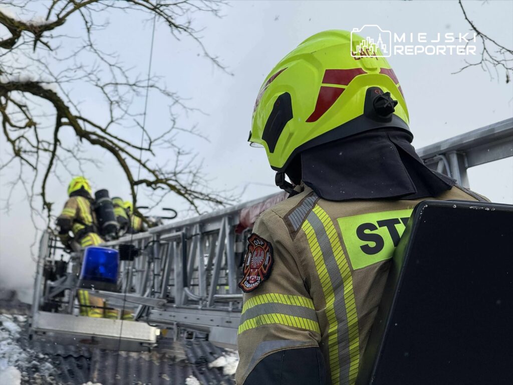 Strażak w żółtej hełmie wspina się po drabinie, podczas akcji gaśniczej w otoczeniu dymu i śniegu.