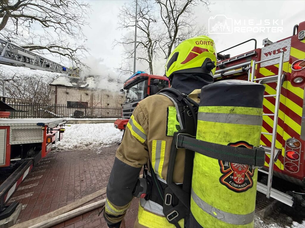 Strażak w specjalistycznym ubraniu obserwuje pożar budynku, z którego wydobywa się dym, w otoczeniu wozów strażackich.