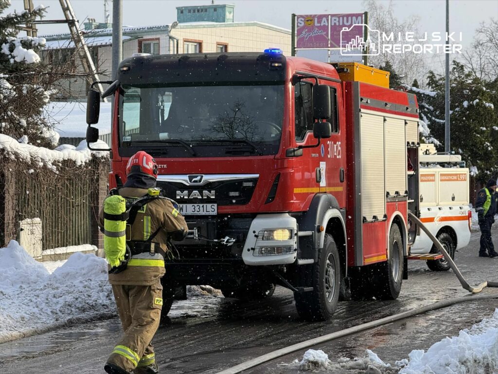 Strażak w hełmie i mundurze zbliża się do wozu strażackiego MAN, obok leży wąż strażacki, a w tle widać zasypane śniegiem budynki.