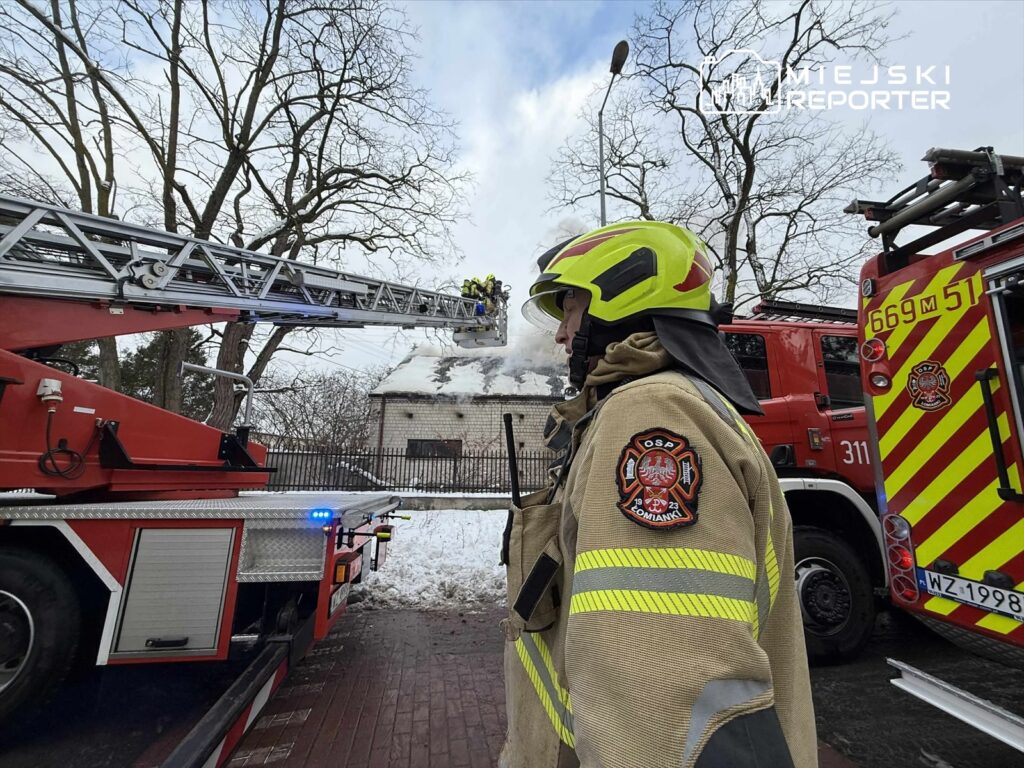Strażak w hełmie obserwujący akcję gaśniczą przy budynku, z którego wydobywa się dym, obok wóz strażacki.