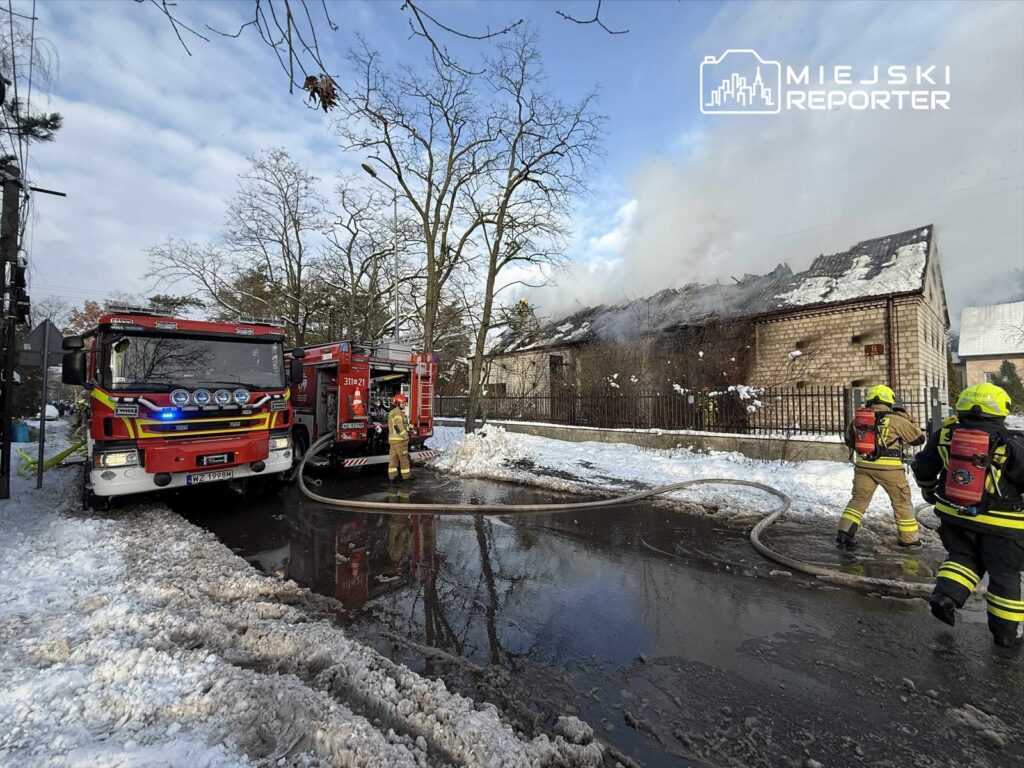 Strażacy z wężami gaśniczymi pracują przy płonącym budynku w zimowej scenerii, otoczeni przez śnieg.