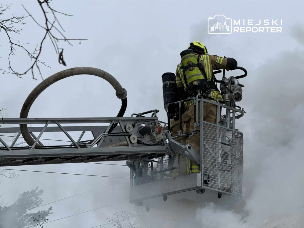 Strażak w specjalistycznym ubraniu obsługuje wąż gaśniczy z podnośnika strażackiego w otoczeniu gęstego dymu.