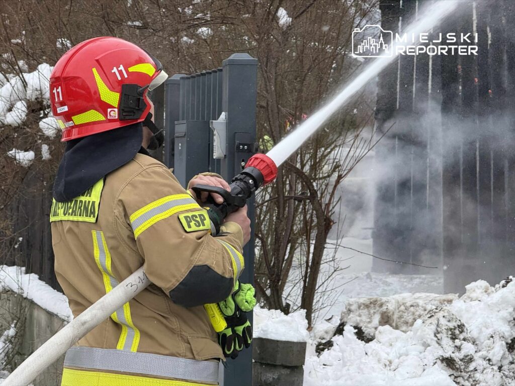 Strażak w hełmie i mundurze gaśniczym używa węża strażackiego do gaszenia ognia w zaśnieżonym otoczeniu.