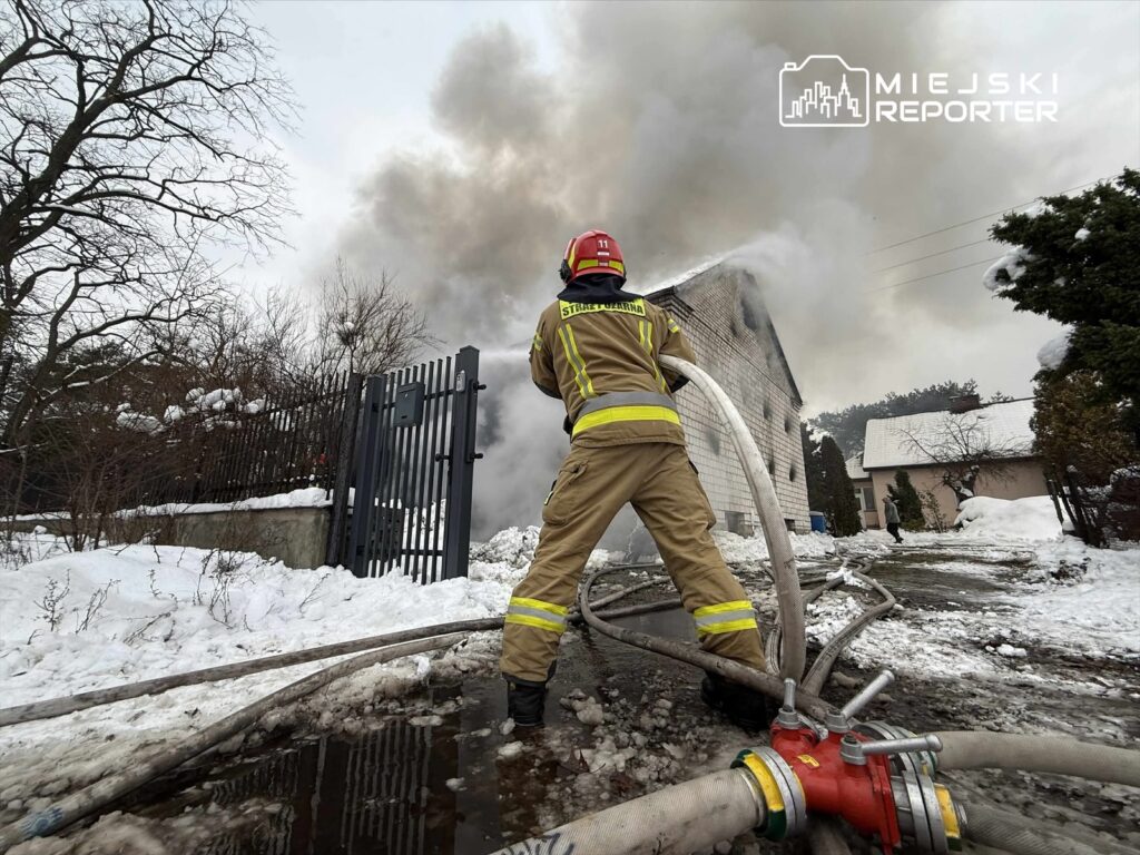 Strażak w hełmie podaje wodę przez wąż, gasząc pożar w domku jednorodzinnym otoczonym śniegiem i dymem.
