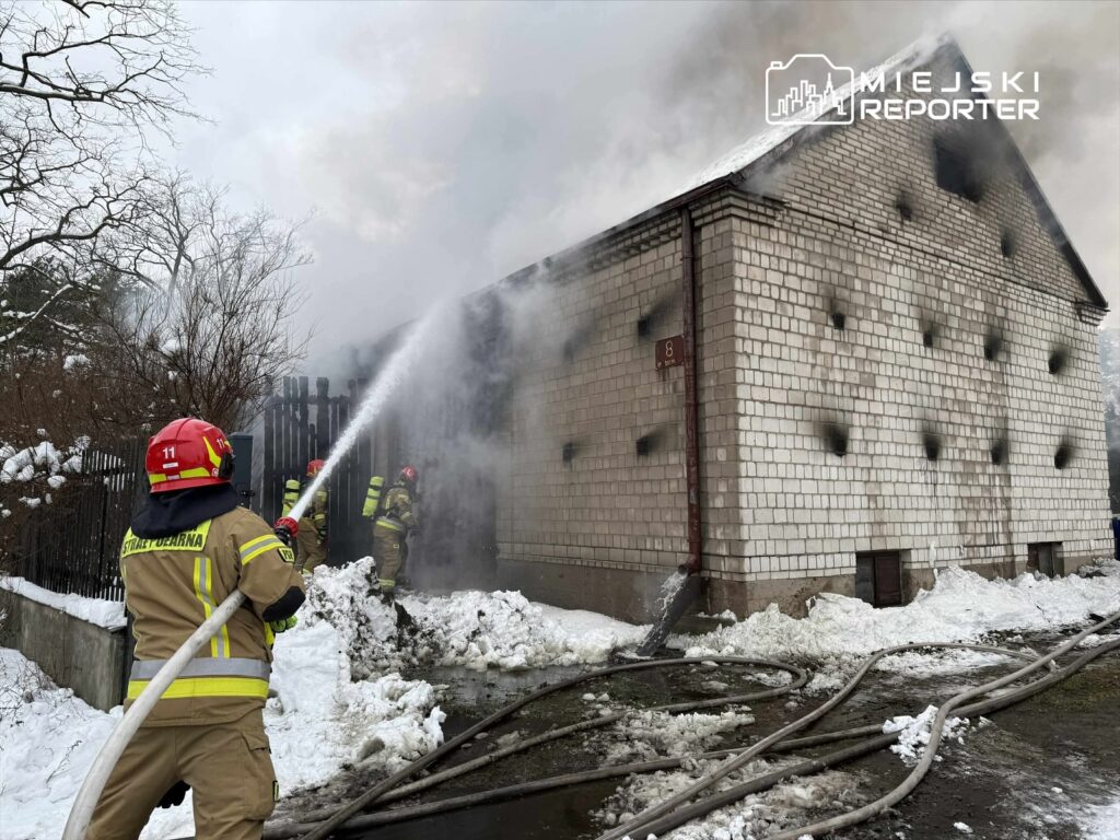 Strażacy w hełmach gaszą pożar budynku mieszkalnego, obok leży śnieg, a w powietrzu unosi się dym.